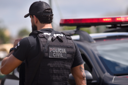 Policial Civil de costas, ao lado de viatura da corporação